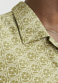 Kurzarmhemd aus einem grünen gemusterten Stoff mit weißen Blumenmotiven. Der Kragen ist strukturiert und hat ein spitzes Design mit auffälligen Nähten.