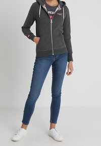 Sweat à capuche zippé gris foncé avec cordons, manches côtelées et poche kangourou, associé à un jean skinny bleu et des baskets blanches.