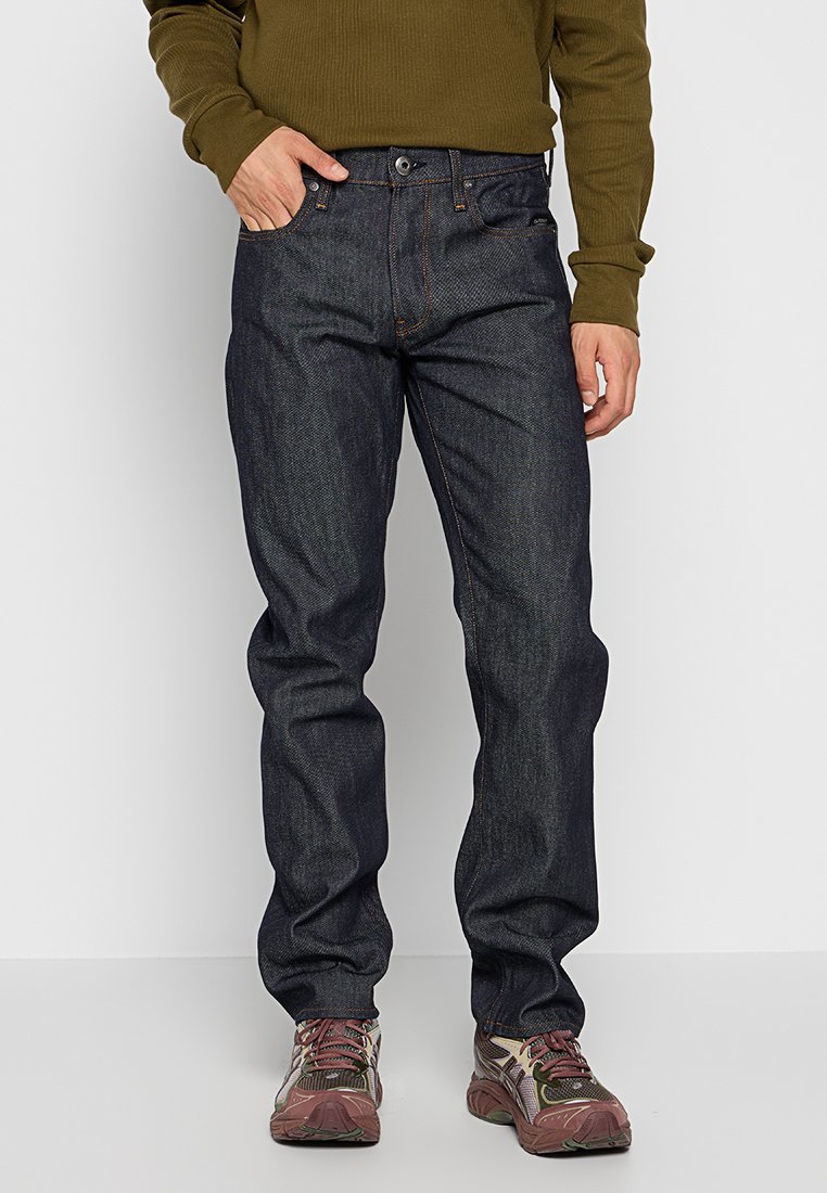 Dunkelblaue Denim-Jeans mit geradem Schnitt, fünf Taschen, Kontrastnähten und mittlerer Leibhöhe, kombiniert mit braunen Sportschuhen.