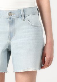 Calções de ganga azul claro com uma bainha desfiada, apresentando um fecho com botão, bolsos frontais e um corte descontraído. Textura suave, design casual.