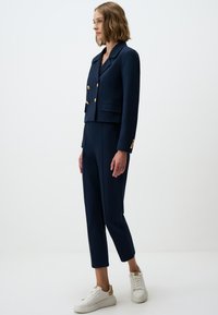 Marineblauwe op maat gemaakte blazer met gouden knopen, gecombineerd met bijpassende slim-fit broek en witte sneakers. Gladde stofstructuur, modern ontwerp.