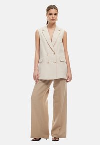 Beige kaksirivinen liivi, jossa on syvä pääntie ja kaulukset, yhdistettynä leveälahkeisiin beigeihin housuihin ja mustiin korollisiin sandaaleihin.