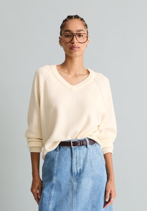 Femme aux cheveux tressés et lunettes portant un pull crème à col en V, rentré dans une jupe en jean bleu taille haute avec une ceinture marron, debout devant un fond uni.