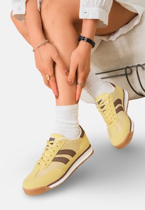 Jambes croisées portant des chaussettes blanches et des baskets jaunes avec des rayures marron, mains reposant sur le bas de la jambe, ornées de bagues et de bracelets.