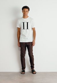 Camiseta blanca con un gran "II" negro impreso en el frente, combinada con pantalones slim-fit marrones y sandalias negras. Textura de tela suave.