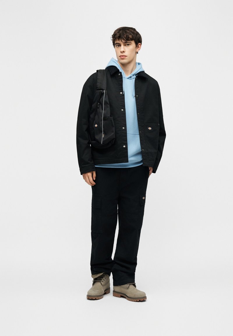 Giovane uomo con indosso una felpa con cappuccio azzurro chiaro, giacca e pantaloni neri, stivali beige e una borsa nera a tracolla, in piedi davanti a uno sfondo bianco.