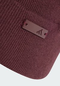 Bordeaux gebreide beanie met een geribbelde textuur en een bruine logo-tag met een drie-strepenontwerp, bevestigd met een bijpassende lus.