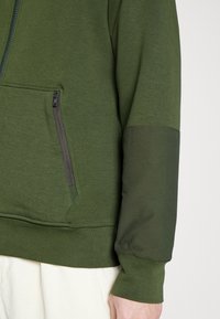 Olivgrön hoodie med halvdragkedja, texturerad tyg, sidoficka med dragkedja och ribbade muddar och fåll.