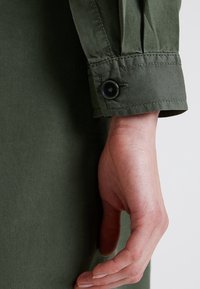 Zelený manžetový knoflík košile s jedním černým knoflíkem, s viditelným prošitím a jemnou strukturou látky.