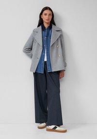 Cappotto in lana grigio doppiopetto sopra una camicia di denim blu, abbinato a pantaloni larghi scuri e sneakers bianche con dettagli neri.