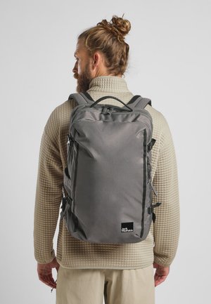 Grauem Rucksack aus strukturiertem Stoff, mit verstellbaren Trägern und Seitentaschen. An der Unterseite befindet sich ein Logo-Tag. Der Rucksack wird von einer Person in einem gesteppte Pullover getragen.