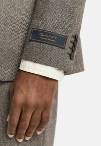 Blazer di tessuto a motivo herringbone marrone con etichetta navy GANT e quattro bottoni neri sui polsini, completato da un polsino di camicia bianca sottostante.