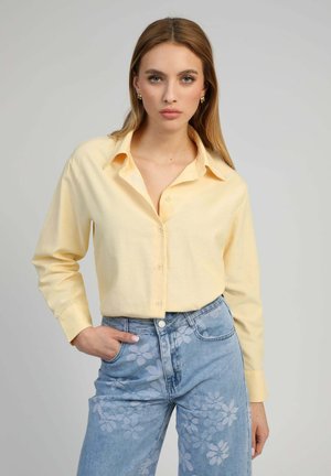 Femme aux cheveux longs portant une chemise boutonnée jaune pâle et un jean taille haute bleu avec des motifs floraux blancs, debout devant un fond uni.