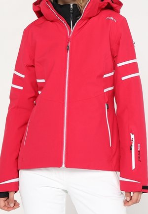 Veste de ski rouge à capuche avec fermeture éclair blanche et bandes réfléchissantes portée sur un pantalon blanc, mettant en valeur des poches zippées et des détails aux manches.