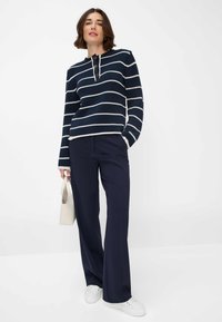 Marineblauw gestreepte trui met witte strepen en vier knopen bij de hals, gecombineerd met marineblauwe broek en witte sneakers. Een lichte handtas wordt vastgehouden.