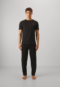 Crna pamučna majica kratkih rukava s malim logotipom na prsima, uparena s odgovarajućim crnim hlačama, prikazana na jednobojnoj pozadini.