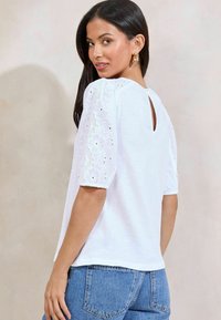 Valge topp lühikeste, punnitatud varrukatega, millel on lilleline silmaorgiga broderie. Kaeluse tagaosa detail, sobitades siniste denimist teksadega.