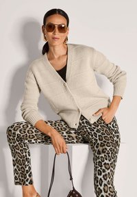 Frau in beigem, knöpfbarem Cardigan, mit Leopardenprint-Hosen, großen Sonnenbrillen und goldenen Ohrringen, sitzt mit einer kleinen braunen Handtasche.