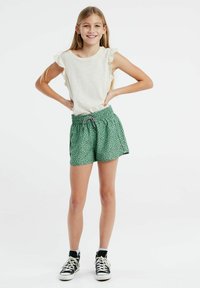 Witte, gerimpelde mouwloze top gecombineerd met groene shorts met een zwart stippenpatroon en een elastische tailleband. Zwarte sneakers maken de outfit compleet.