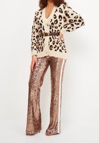 Cardigan con stampa leopardata in crema e nero, indossato sopra una maglietta bianca, abbinato a pantaloni bronzo a vita alta con paillettes e bande laterali bianche.