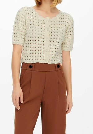 Femme portant un haut en crochet crème à manches courtes et boutonné, associé à un pantalon taille haute marron avec des boutons noirs.