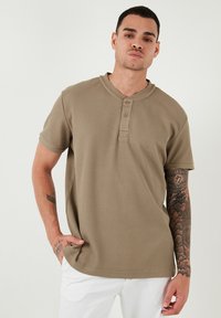 Kaki színű, rövid ujjú henley póló, texturált anyagból készült, három fakerettel, kerek nyakkivágással és laza szabással.