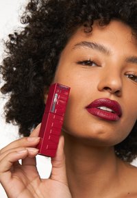 Mörkröd läppstift i en elegant, rektangulär tub med en texturerad yta och präglad logotyp. Modell visar produkten på en jämn hy.