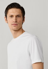 Junger Mann mit kurzen dunklen Haaren, der ein schlichtes weißes T-Shirt trägt und leicht seitlich vor einem hellgrauen Hintergrund steht.