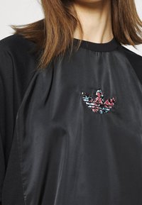 Detailní záběr na černou košili s lesklým předním panelem a květinovým vzorem loga Adidas trefoil na prsou.