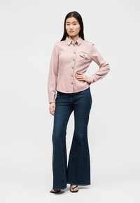 Kvinne står med en hånd på hoften og har på seg en lyserosa skjorte med knapper og mørkeblå vide jeans med svarte sandaler.