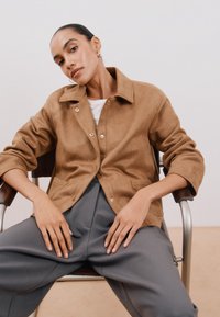 Ruskea mokkajakku, jossa on kauluskietaisu, nepparipainikkeet ja etupocketit, yhdistettynä harmaisiin leveälahkeisiin housuihin. Malli istuu tuolilla.