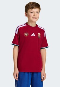 Červený športový dres s bielymi a zelenými akcentmi, s maďarským erbom a kruhovou známkou. Vyrobený z priedušnej tkaniny, s okrúhlym výstrihom.