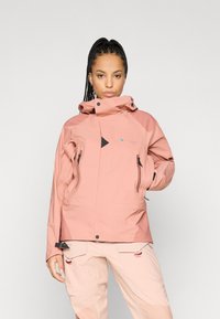 Casaco rosa impermeável com gola alta, apresentando realces pretos, bolsos com fecho e um design geométrico. Usado sobre calças de cor clara.