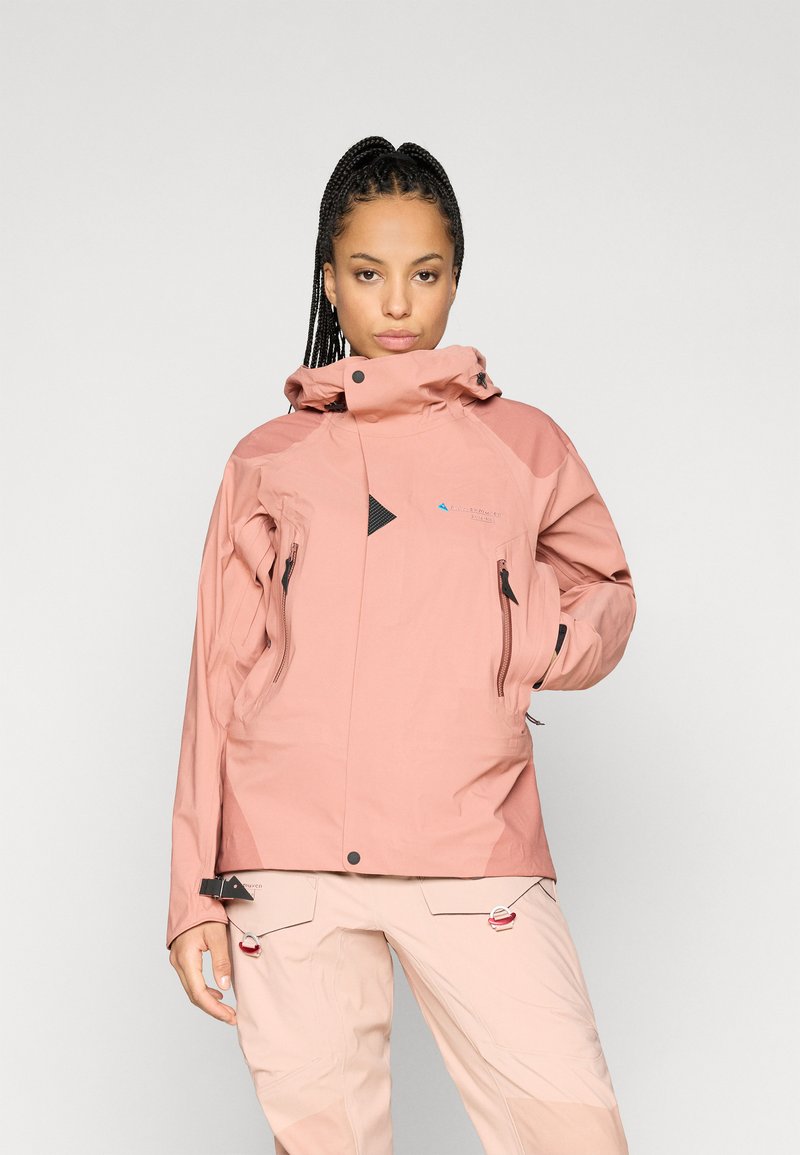 Casaco rosa impermeável com gola alta, apresentando realces pretos, bolsos com fecho e um design geométrico. Usado sobre calças de cor clara.