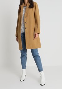 Kamelhaar-Mantel mit einreihigem Design, ausgestattet mit Knöpfen, Seitentaschen und einer maßgeschneiderten Passform, kombiniert mit blauen Jeans und weißen Stiefeletten.