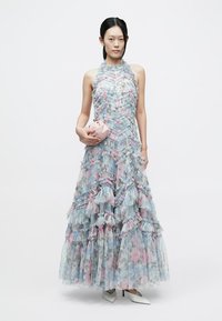 Lichtblauwe bloemenjurk met laagjes en ruches, halternek en doorschijnende textuur. Gecombineerd met een kleine roze clutch en witte schoenen met hoge hakken.