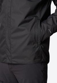 Personne portant une veste imperméable noire avec une main dans la poche, texte « "DRYVENT" » visible sur la manche, sur fond uni.