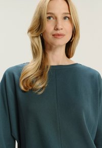 Blondynka z luźnymi włosami, nosząca złote kolczyki w kształcie obręczy i teksturowaną bluzkę w kolorze turkusowym, patrząca nieco poza kamerą na jednolitym jasnym tle.
