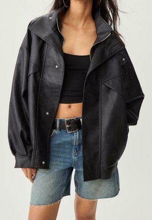 Femme portant une veste en cuir noire oversize sur un crop top noir et un short en denim effiloché avec une ceinture noire et une boucle argentée.