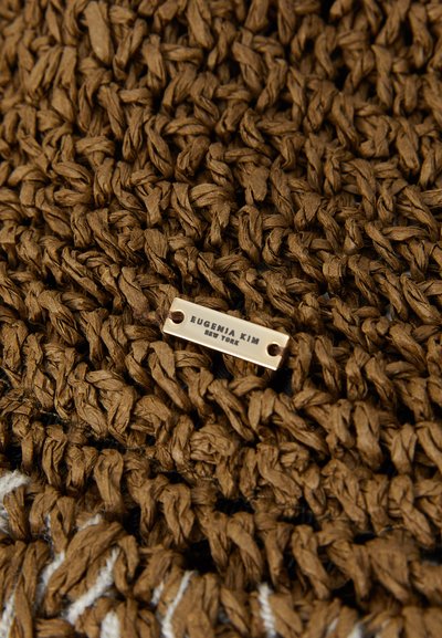 Barna szőtt textil, textúrált szálakkal és egy fém címkével, melyen a felirat: "EUGENIA KIM NEW YORK." A mintázatban összefonódó szálak láthatók.