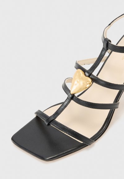 Sandalia de cuero negro con punta cuadrada, múltiples correas delgadas y un acento en forma de corazón dorado en la parte superior. Textura suave, diseño minimalista.