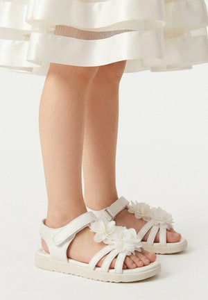 Kind trägt weiße Sandalen mit drei Riemen und dekorativen Blumenapplikationen, kombiniert mit einem weißen Kleid mit transparenten horizontalen Streifen.