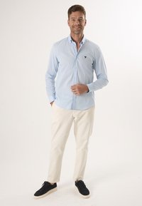 Helles, blaugraues Hemd mit einer kleinen dunklen Logo, getragen mit weißen Hosen und dunklen Sneakers. Das Hemd hat einen spitzen Kragen und lange Ärmel.