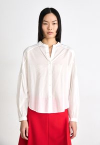 White, long-sleeve button-up shirt made of lightweight fabric features a relaxed fit, rounded hem, and subtle texture, paired with a red skirt.