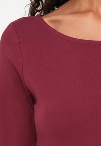 Burgundi hosszú ujjú felső, széles nyakkivágással, sima, rugalmas anyagból készült; illeszkedő szabású és minimális varrásokkal rendelkezik.