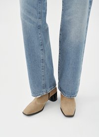 Beige suede ankle boots with a pointed toe and a low black heel, worn with light blue straight-leg jeans featuring frayed hems.