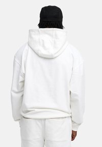 White hooded sweatshirt with a relaxed fit, large hood, and long sleeves. Made from soft fabric, features ribbed cuffs and hem, and is paired with white pants.
