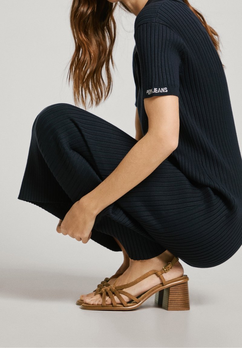 Robe bleu marine côtelée à manches courtes avec un logo sur le bras, associée à des sandales à talons hauts marron clair à lanières et talon carré.