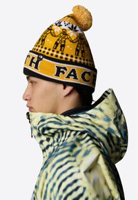 Włóczkowa czapka beanie w kolorze żółtym z czarno-białymi wzorami, z pomponem na czubku i logo "THE NORTH FACE" na pasku.
