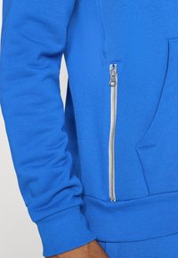 Blauer Sweatshirt mit einer Reißverschlusstasche auf der linken Seite, gerippten Bündchen und glatter Textur. Das Material wirkt weich und leicht.